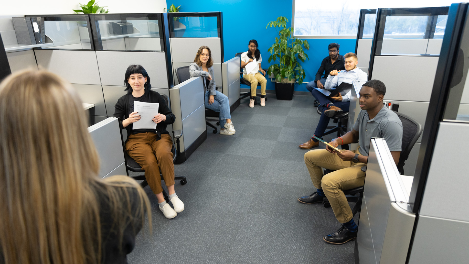 A group of staff sits at the edge of their pods to look at a blonde woman speaking in front of them at the Bioscience and Technology Business Incubator in Lawrence, KS.
