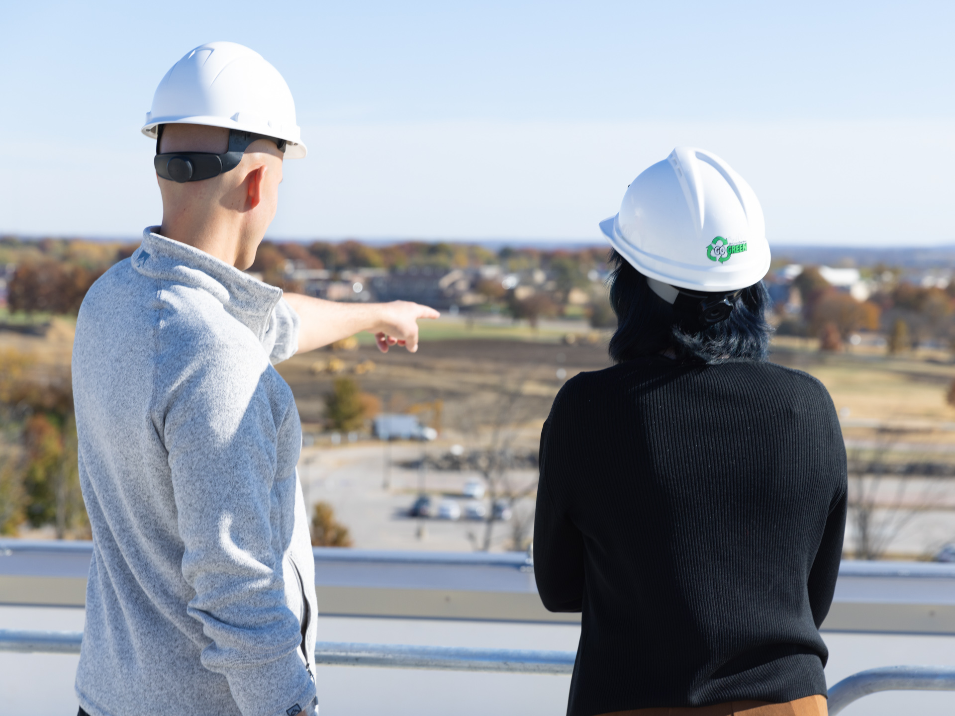 Two people in hardhats stand on a roof and point to land below while furthering the economic development of Lawrence.
